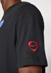 Sort T-shirt med korte ærmer, der har et blåt grafik på brystet og et rødt broderet Nike-logo på venstre ærme. Blødt bomuldsstof.