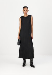 Rochie neagră fără mâneci, cu textură plisată, siluetă dreaptă și decolteu rotund. Purttată cu cizme negre cu toc și o poșetă mică.