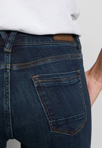 Ciemnoniebieskie dżinsy z pomarańczowym szwem, z małą tylną kieszenią i skórzaną naszywką przy pasku. Zbliżenie z tyłu.