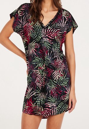 Kobieta w sukience z krótkim rękawem i dekoltem w serek, w liściasty wzór w kolorach czerwonym, różowym, zielonym i czarnym, z regulowanym taliowaniem na troczki zakończone frędzlami.