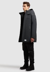 Czarna ocieplana kurtka z wysokim kołnierzem i kapturem, z zamykanymi na zamek kieszeniami i naszywką z logo, noszona na czarnych spodniach cargo i botkach.