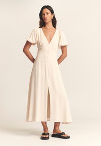 Lichtbeige maxi-jurk met een diepe V-hals, korte pofmouwen, knoopsluiting aan de voorkant en een split. Draagt zwarte platte sandalen.