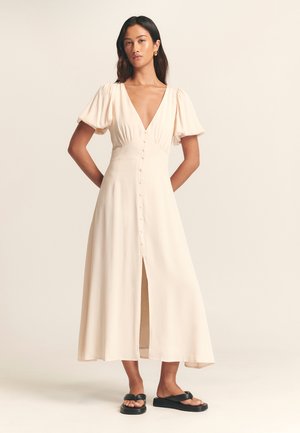 Vaaleanbeige midi-mekko puff-hihnoilla, syvä V-kaula-aukko, etupainonappikiinnitys ja sivuventtiili. Yhdistetty mustiin korokepolkupöksyihin.