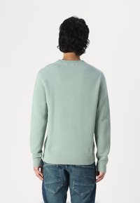 Suéter de malha verde claro com gola redonda canelada, mangas longas e padrão texturizado, combinado com jeans de ganga, visto por trás.