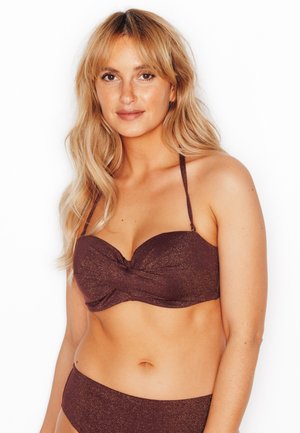 Femme aux cheveux longs blonds portant un maillot de bain deux pièces marron foncé scintillant, souriante sur un fond blanc.