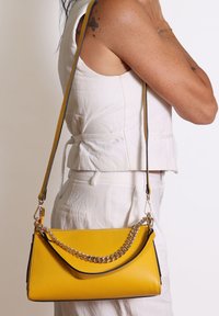Borsa a tracolla in pelle gialla con finitura texture, caratterizzata da un accento in catena e una tracolla a contrasto. Forma rettangolare con profilo sottile.