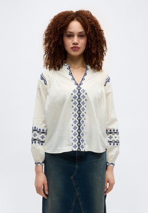 Femme aux cheveux bouclés portant un chemisier blanc avec une broderie bleue et une jupe en jean foncé, face à la caméra sur un fond uni.