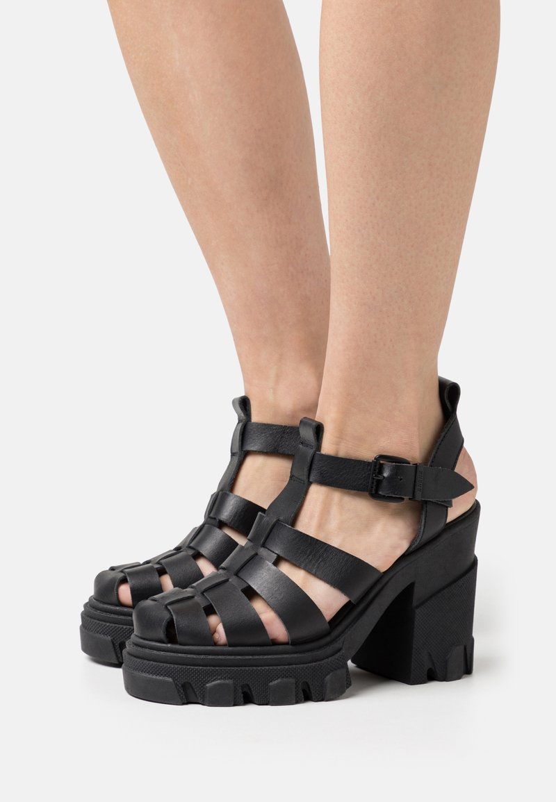 Sandales noires à plateforme avec un talon épais, présentant un design en cuir à lanières et une fermeture à boucle. Les semelles texturées offrent une bonne adhérence.