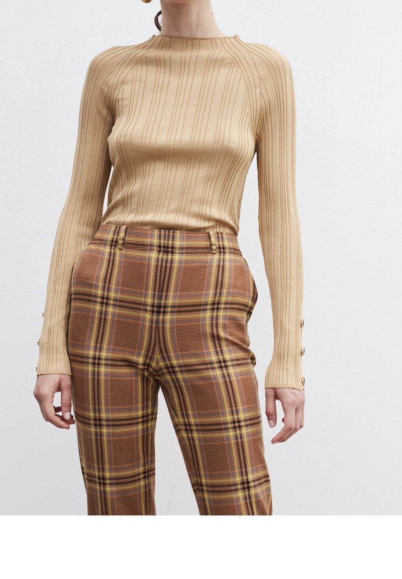 Femme portant un haut beige côtelé à manches longues avec des détails de boutons et un pantalon marron à carreaux taille haute sur fond blanc.