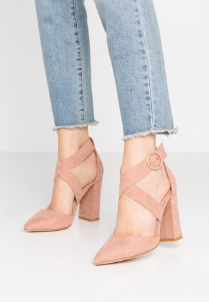 Różowe zamszowe szpilki z krzyżującymi się paskami wokół kostki i okrągłą klamrą. Charakteryzują się spiczastym czubkiem i masywnymi obcasami z pionowymi żłobieniami.