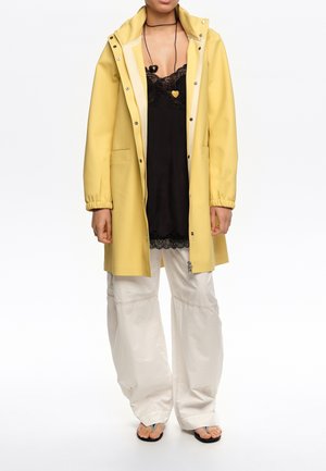 Henkilö pukeutunut keltaiseen hupulliseen sadeviittaan mustan pitsireunaisen yläosan päällä, valkoisiin leveälahkeisiin housuihin, mustiin varvassandaaleihin ja kahteen sydämenmuotoiseen kaulakoruun.