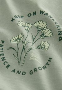 Šviesiai žalia medžiaga su botaniniu raštu, kuriame pavaizduoti stilizuoti balti gėlės ir tamsiai žaliais tekstais "Tęsk keliones" ir "Kantrybė ir augimas."