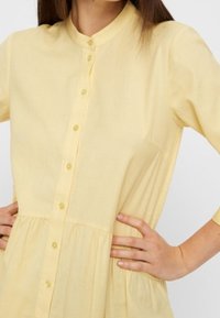 Gelbes Hemd mit Stehkragen, Taillenraffung und weichem, leichtem Stoff. Dreiviertelärmel.