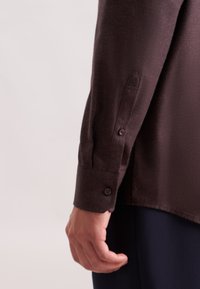 Camicia marrone a maniche lunghe con una texture liscia, dotata di polsini con bottone e un fit leggermente rilassato. Inquadratura ravvicinata che mostra la manica e la mano.