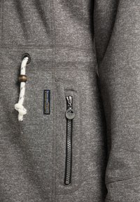 Šedá látka mikiny s texturovaným povrchem, kapsou na zip s černým lemováním a bílým stahovacím jezdcem s dřevěnými korálky.