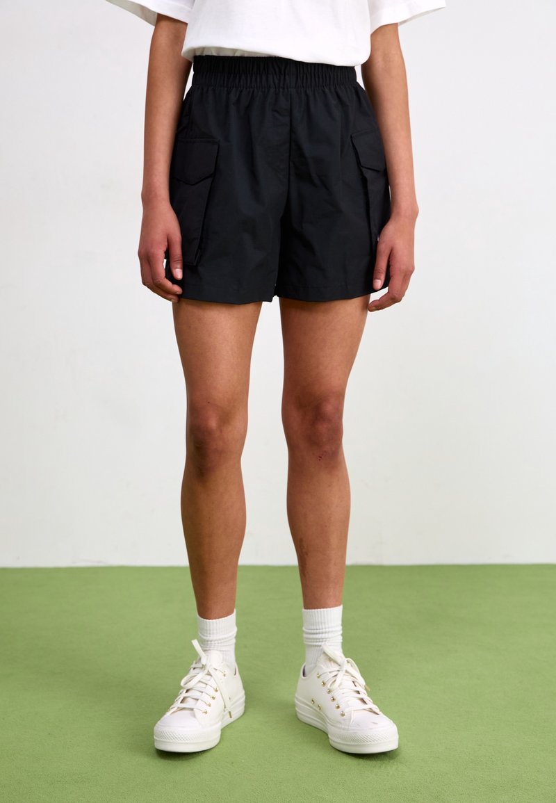 Black cargo shorts with an elastic waistband, featuring two side pockets and a lightweight fabric. Worn with white sneakers and socks.