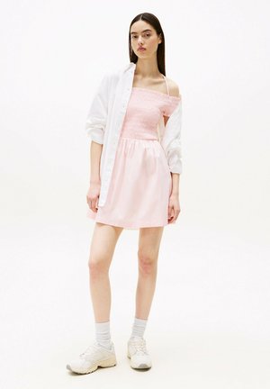 Robe rose clair avec un corsage smocké et une jupe évasée, associée à une chemise blanche oversize et des chaussures de sport beiges.