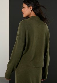 Oliwkowy, oversize sweter z okrągłym dekoltem, długim rękawem i teksturowanym materiałem. Ma opadające ramiona i luźny krój.