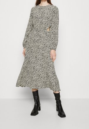 Kobieta ubrana w czarną i białą kwiatową midi sukienkę z długim rękawem i bocznym wycięciem, w połączeniu z czarnymi botkami na obcasie o kwadratowym czubku.