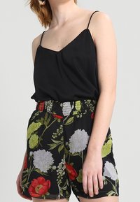 Camisola nera con spalline sottili abbinata a pantaloni shorts a vita alta con un vivace motivo floreale in rosso, bianco e verde su uno sfondo nero.