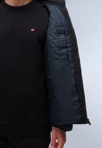 Svart sweatshirt med en liten röd och blå flagga, bärs under en svart puffjacka med slät marinblå foder och elastiska muddar.