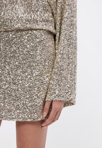 Zlata obleka s kroglicami, ohlapnega kroja in dolgimi rokavi, ki se ponaša s sijočo teksturo in robom, ki sega nad kolena.