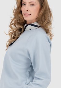 Felpa azzurra con scollo a girocollo e maniche raglan. Una sciarpa a motivi blu navy e bianca è appoggiata intorno al collo. Texture morbida.