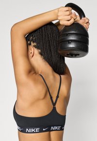 Melna sporta krūšturis ar plānām lencītēm un Nike logotipa joslu. Persona tur melnu kettlebelli virs galvas, demonstrējot tā reljefēto virsmu.