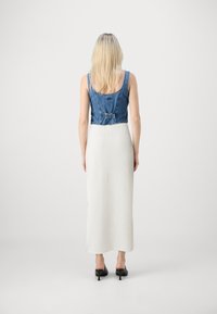 Denim crop top z nastavljivim hrbtom, kombiniran z dolgim, rebrastim belim krilom in črnimi visokimi petami. Nevtralna ozadje.