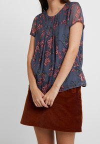 Blus med blommönster och korta ärmar i blått tyg, med fickor och avslappnad passform, ihop med en brun kordflod mini kjol.