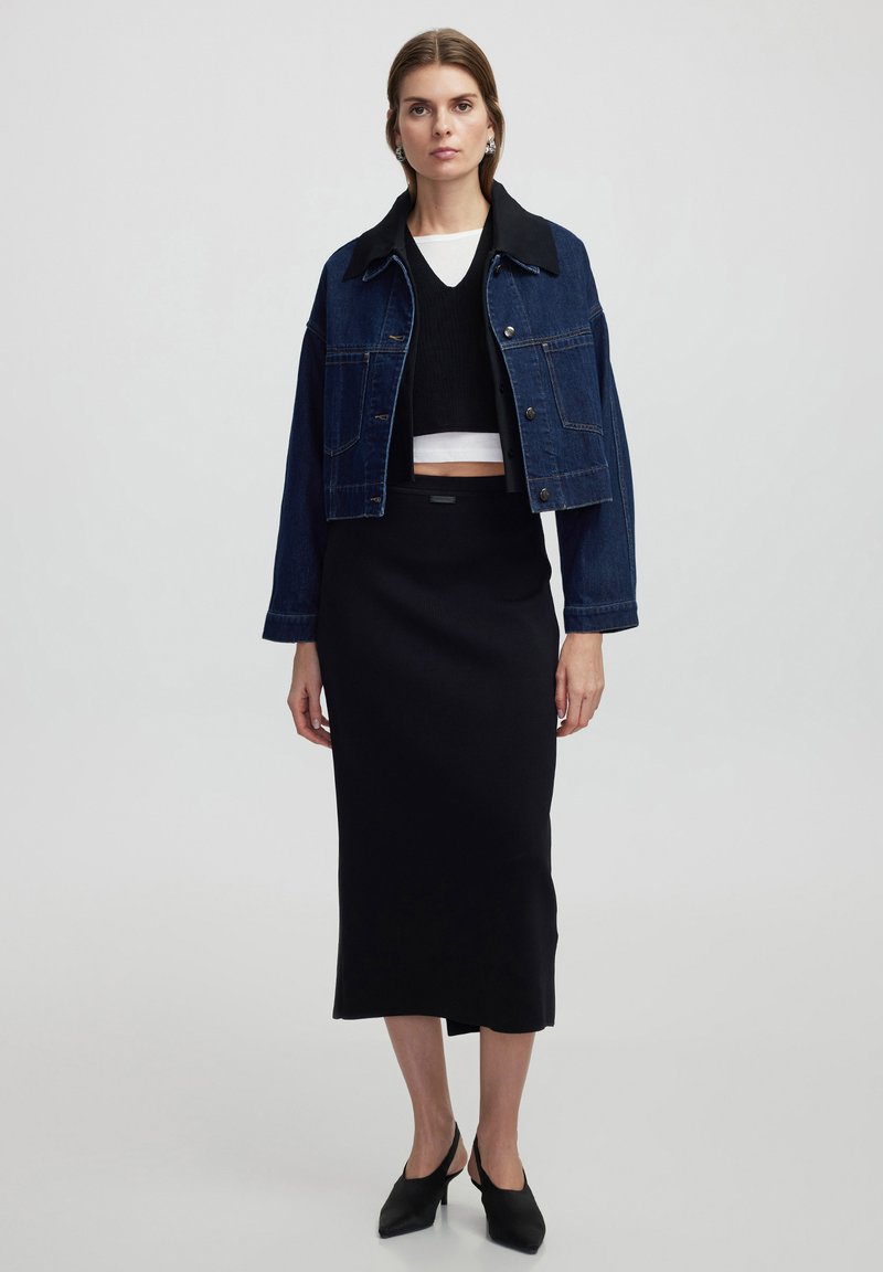 Chaqueta de mezclilla con cuello negro, suéter negro corto y falda midi negra. La modelo lleva zapatos de tacón negro de punta. Diseño sencillo y ajustado.