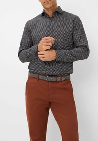 Schwarz gemusterte Bluse mit kleinen weißen und goldenen geometrischen Formen, lange Ärmel, geknöpfter Kragen; kombiniert mit braunen Hosen und einem grauen Gürtel.