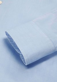 Puño de camisa de manga larga azul claro con costuras visibles y una textura sutil, doblado de manera ordenada sobre un fondo de tela a juego.