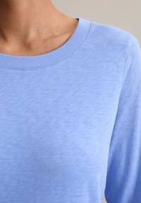 Nahaufnahme einer Person, die ein hellblaues Rundhalsstrickoberteil trägt, wobei der Ausschnitt, die Schulter und der obere Brustbereich sichtbar sind.