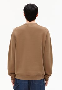 Persoon met kort zwart haar die wegkijkt, rondgeleid in een bruine longsleeve sweatshirt en donkerblauwe broek met grote achterzakken.