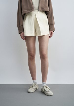 Person iført cremefarvede plisserede shorts, beige lynlåsjakke, hvide sokker og beige sneakers, står på gråt gulv mod en lys væg.