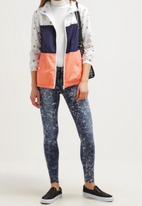 Farveblokkeret jakke med marineblå, koral og hvide mønstrede ærmer. Tætsiddende jeans i et slidteblåt mønster og sorte slip-ons.