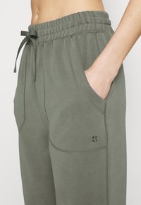 Pantaloni della tuta verde-grigio con vita elastica e coulisse, dotati di tasche laterali e un piccolo dettaglio con logo sulla coscia.