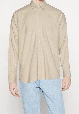 Chemise beige à manches longues avec boutons et poche poitrine portée avec un jean bleu clair sur une personne debout, cadrée au niveau du cou et des cuisses.