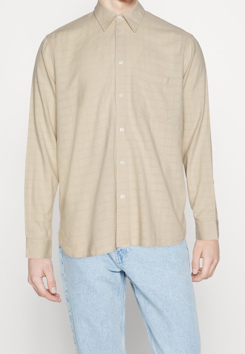 Chemise beige à manches longues avec boutons et poche poitrine portée avec un jean bleu clair sur une personne debout, cadrée au niveau du cou et des cuisses.