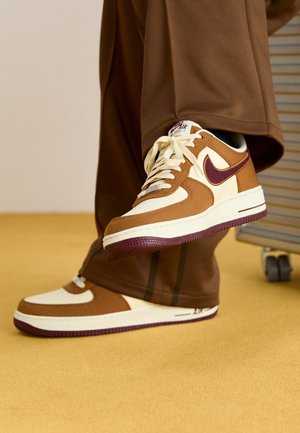 Brązowe i kremowe sneakersy Nike Air Force 1 noszone z brązowymi spodniami na żółtej podłodze obok krzesła na kółkach.