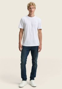 Balti medvilninė palaidinė su apvalia iškirpte, derinama su tamsiai mėlynais džinsais ir baltais sportbačiais. Modelis stovi neutraliame fone.