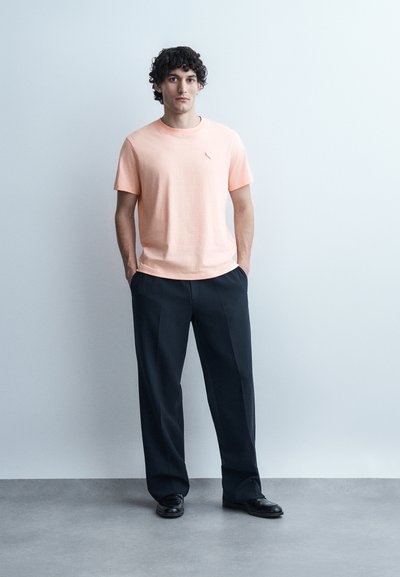 Άνδρας με σγουρά μαλλιά να στέκεται, φορώντας ένα ανοιχτό ροζ μπλουζάκι, μαύρο παντελόνι και μαύρα παπούτσια, μπροστά σε απλό ανοιχτό φόντο.