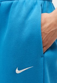 Χέρι που ακουμπά στην τσέπη ενός μπλε παντελονιού Nike με εμφανές λευκό λογότυπο Nike swoosh στο ύφασμα.