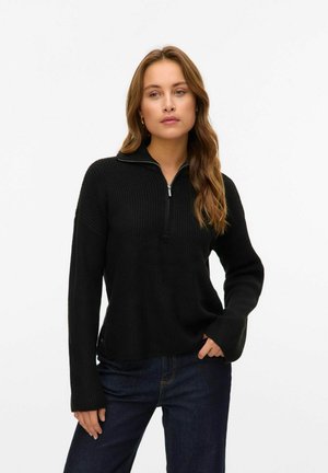 Pull en tricot noir avec un col half-zip, texture côtelée, manches longues et coupe ample, associé à un jean en denim foncé.