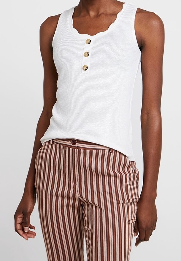 Femme portant un haut blanc côtelé sans manches avec trois boutons et un pantalon marron rayé à taille haute, debout les bras détendus.
