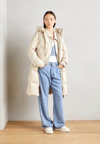 Beige pufferjack met capuchon, denim rompertje en lichtblauwe wijde jeans. Witte sneakers met textuurdetails. Minimalistisch ontwerp.
