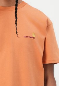 Korte mouwen oranje katoenen t-shirt met een logo op de linkerborst in paars en geel. Soepele textuur met een geribbelde ronde hals.