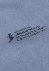 Svijetloplavi materijal s crnim tekstom: "Indigofera tinctoria Cvijeće za ljude. Ovaj jedinstveni komad obojen je isključivo cvijećem i mineralima."