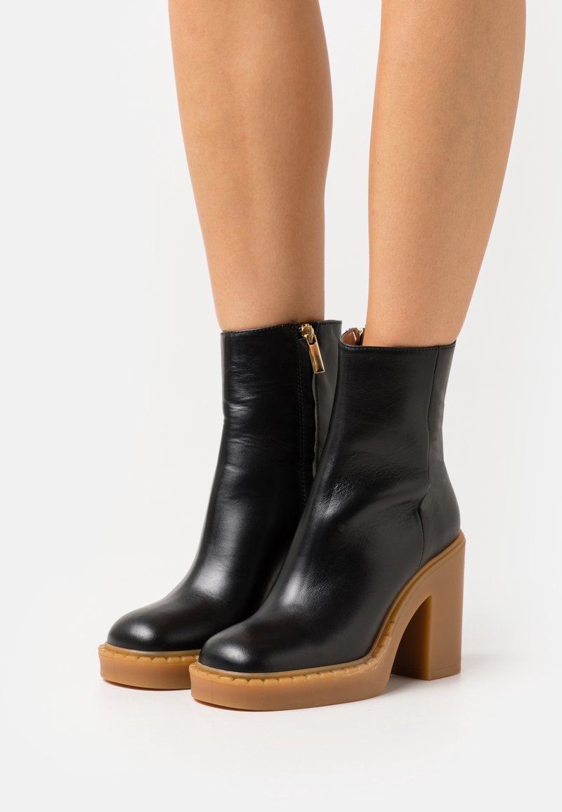 Botines de cuero negro con un tacón alto de bloque, punta redondeada, cremallera lateral y una suela plataforma en tono marrón claro. Textura lisa con un acabado pulido.
